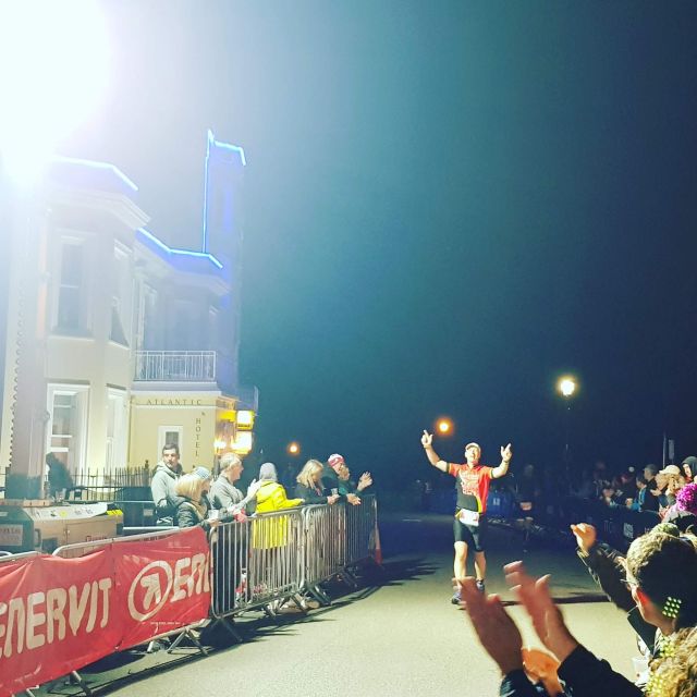 A man runs towards the finish line of Ironman Tenby, his arms are aloft and he's smiling at the camera. People line the street cheering