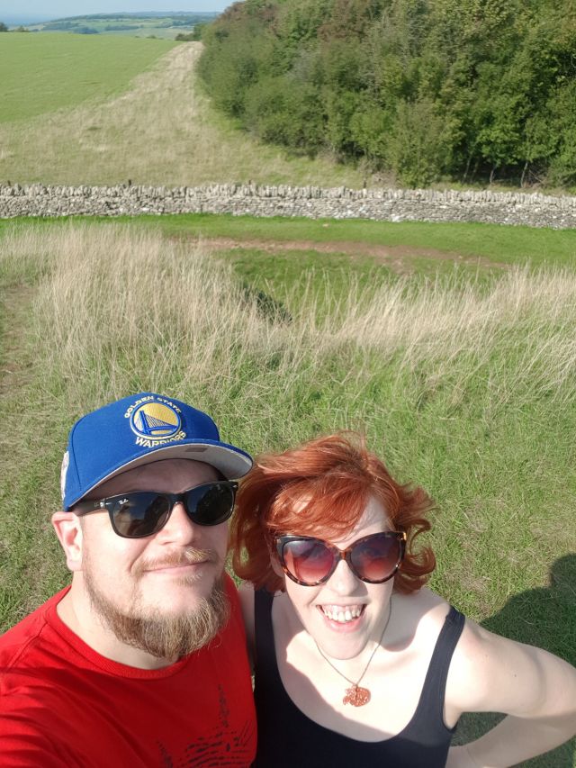 A man and a woman stand on top of a neolithic long barrow (a grassy hill) smiling at the camera