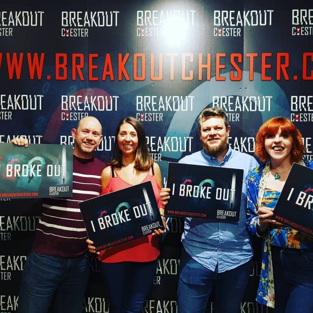 Four people, two men and two women stand in front of a wall advertising "Breakout Chester". They are all smiling and holding signs that say "I broke out"