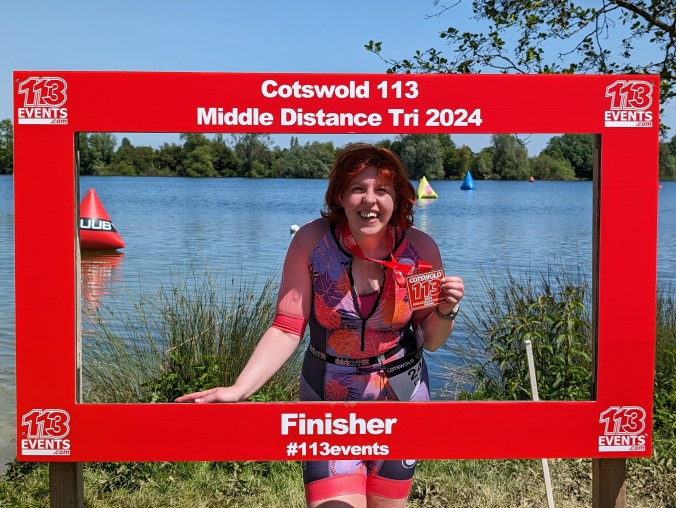 A white woman with red hair smiles broadly as she holds up a large red Cotswold 113 2024 Middle Distance Triathlon medal. She's wearing a pink and purple triathlon suit and standing in a red frame in front of a lake