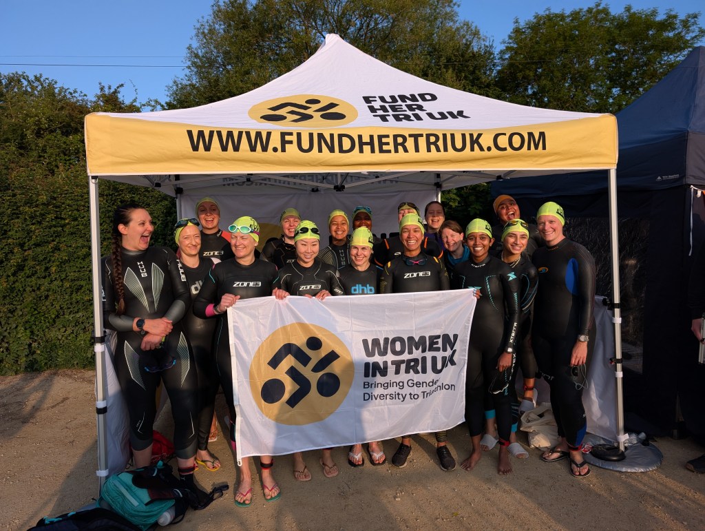 A group of women wearing wetsuits and swimming hats smile for the camera. They're standing under a white and yellow gazebo with the name and logo "Fund Her Tri UK" printed on it.
