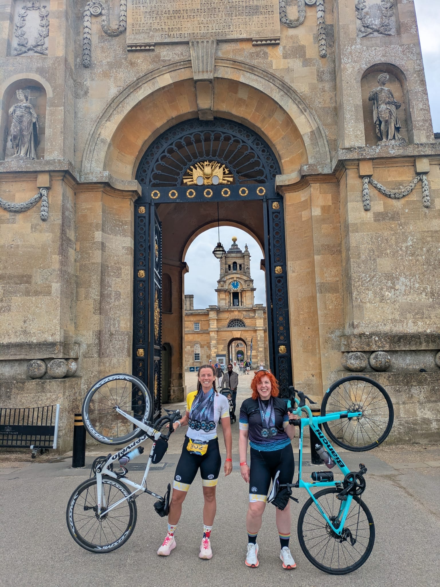 Two women standing in front of the gates of Blenheim Palace, both are holding their bikes next to them and smiling at the camera with their medals around their necks.