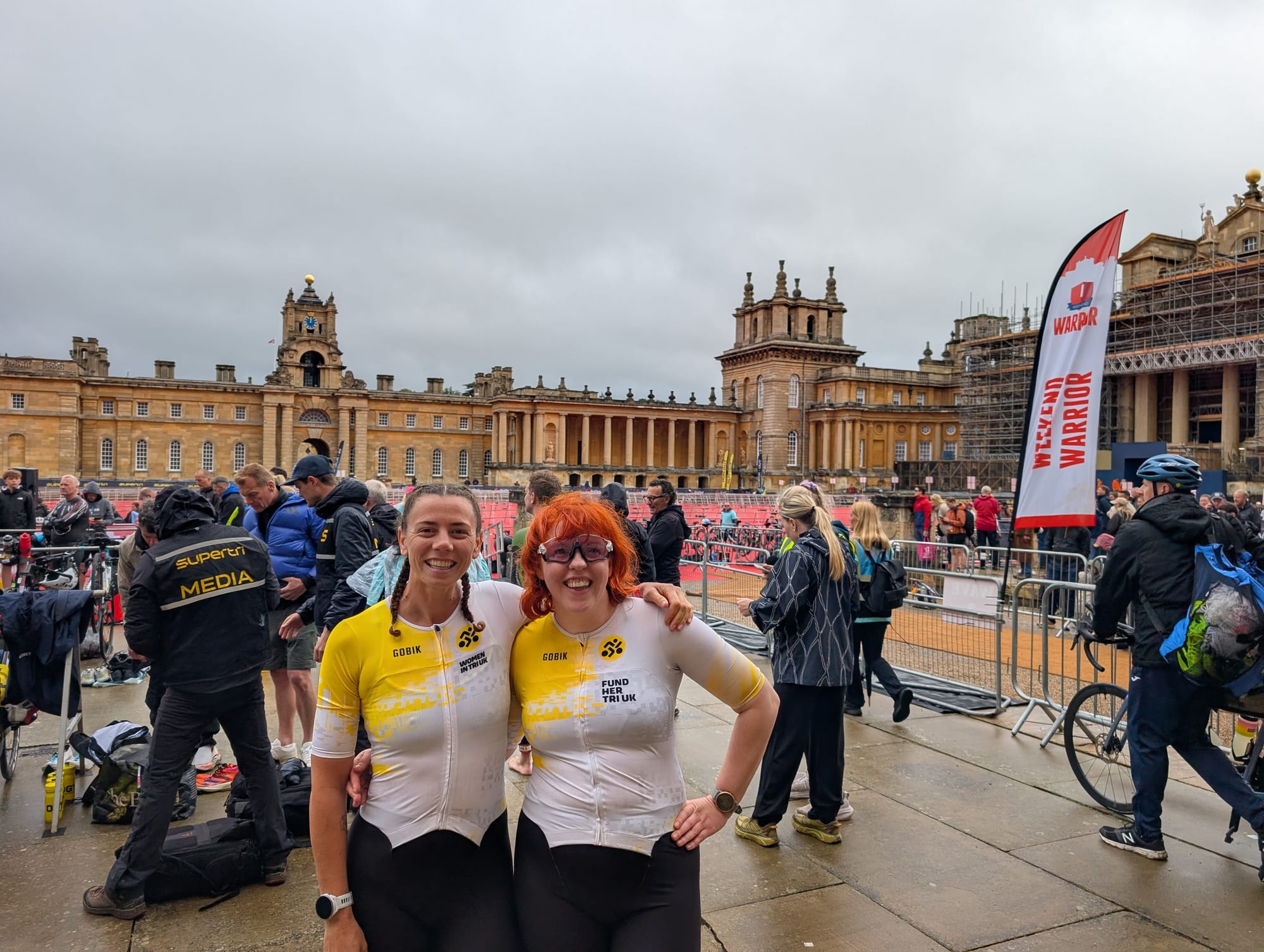 Two women in Women in Tri UK trisuits standing smiling in the courtyard of Blenheim Palace. There are a group of people behind them taking pictures of Jonny Brownlee's triathlon kit