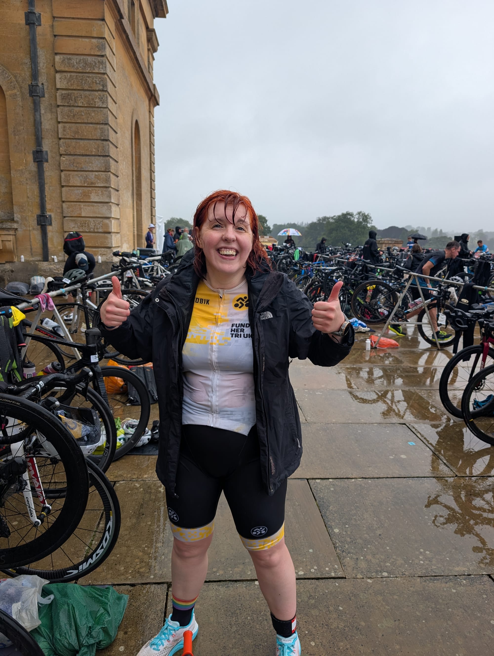 Me, wearing a yellow and black trisuit under a black raincoat, smiling and putting two thumbs up to the camera despite being absolutely drenched through
