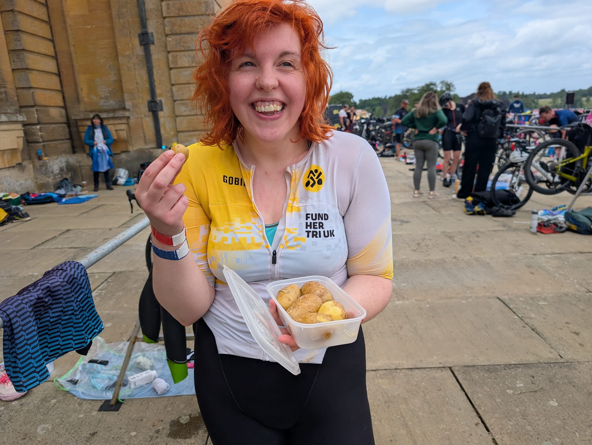 Me standing in the transition area wearing a yellow and black women in tri uk trisuit and eating new potatoes out of a tupperware box. I look very happy about the whole situation!