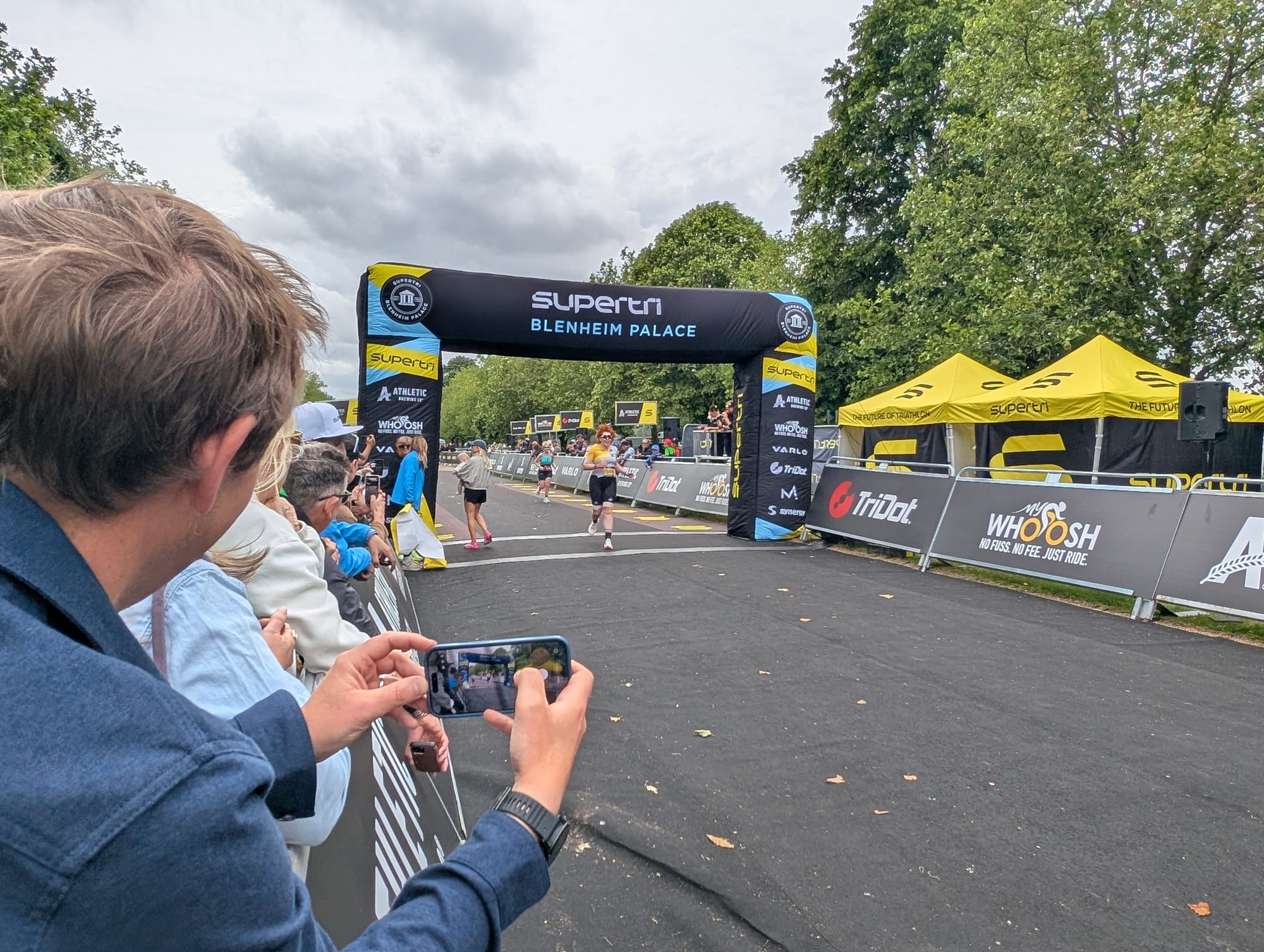 Me wearing my yellow and black trisuit and running under a black finish gantry covered in Supertri Blenheim Palace branding. On the left hand side of the photo you can see a blonde woman holding a baby - she's Jonny Brownlee's wife waiting for him to finish.